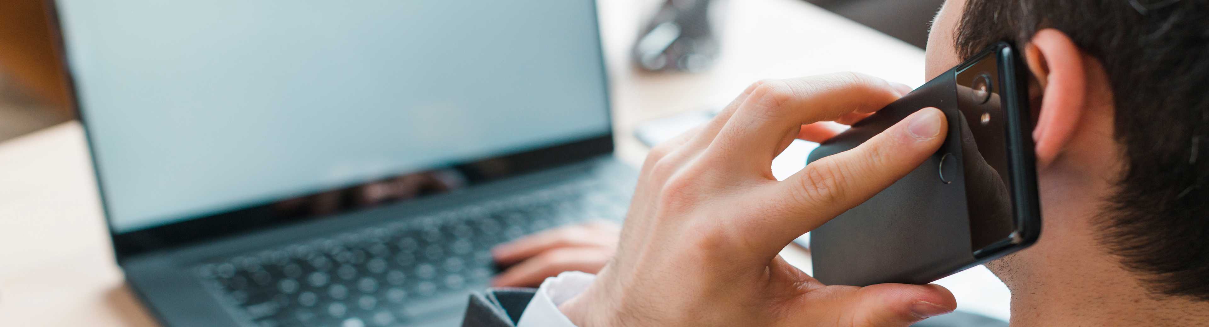 A man converses on a cell phone while seated at a desk with a laptop open in front of him.