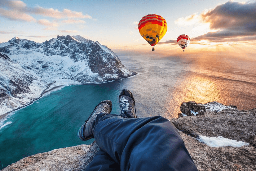 A person sits on a rock, gazing at the ocean while colorful hot air balloons float in the sky above.