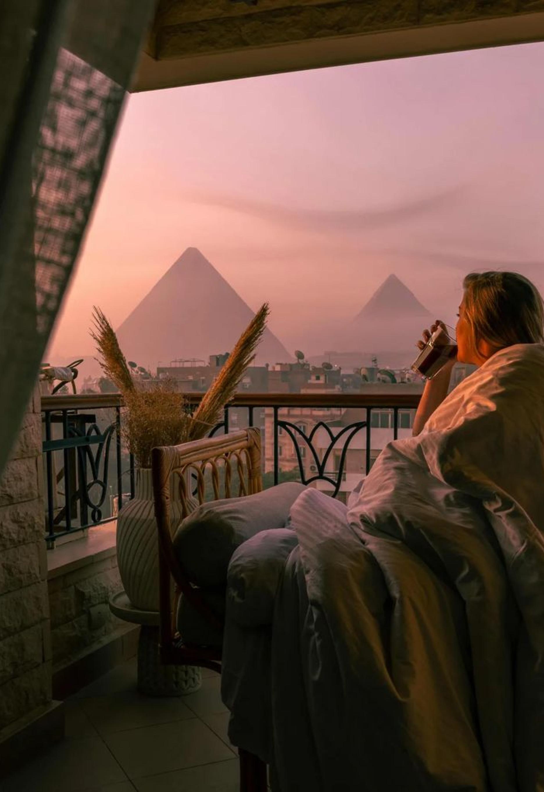 A woman enjoys coffee in bed, with the pyramids prominently displayed outside her window.