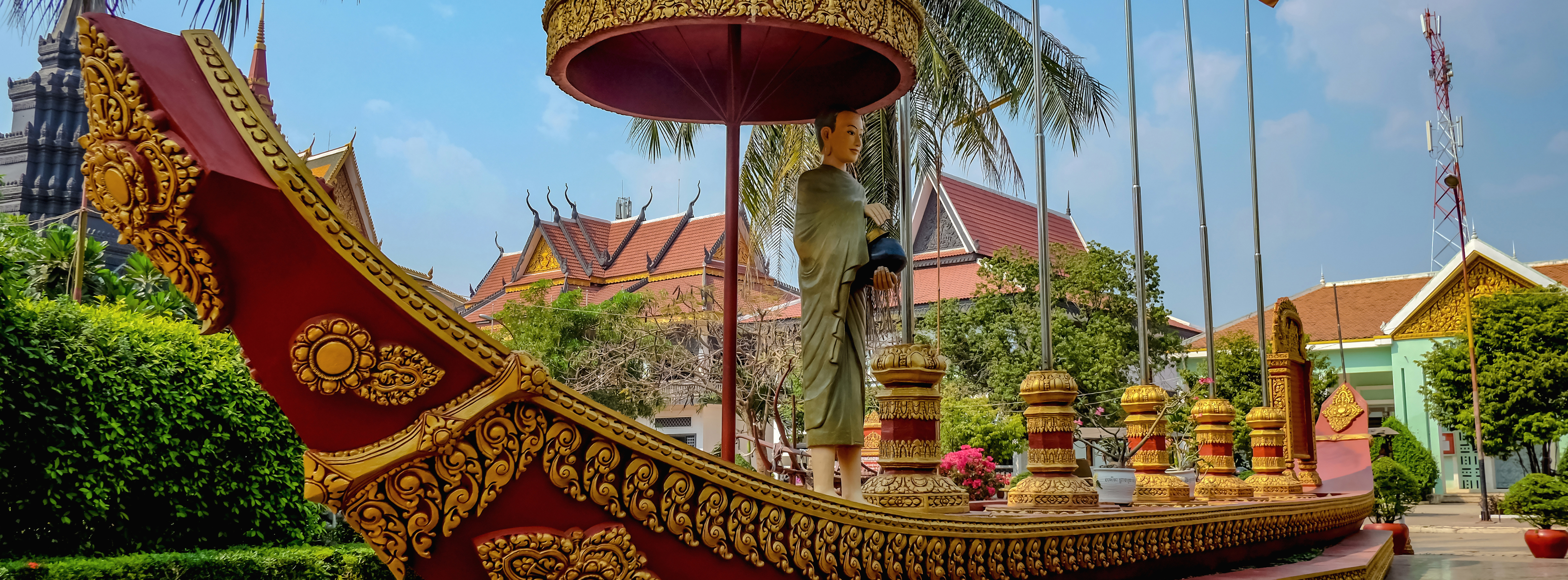 Ornate red and gold boat-shaped monument in front of a traditional Southeast Asian building, featuring a statue of a person in a robe.